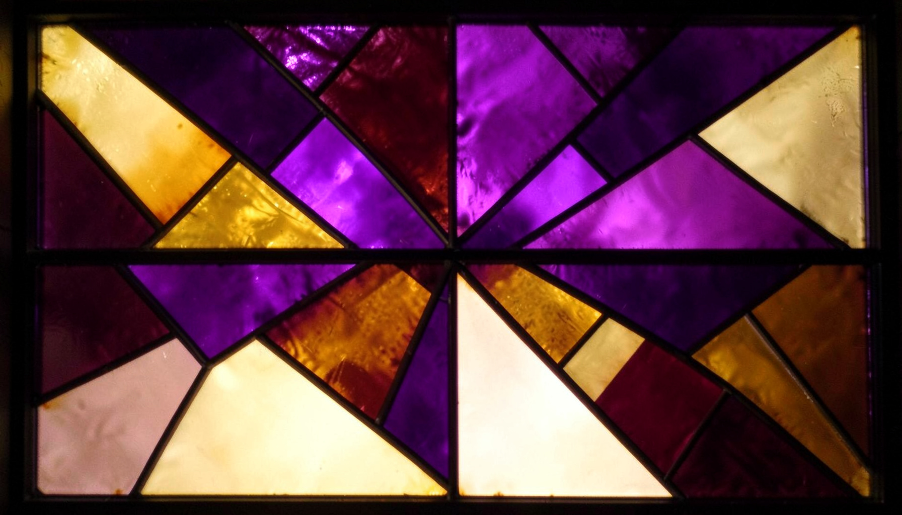 A stained-glass composition with bold leaded segments in deep plum, amber gold, and soft cream tones, forming an abstract luminous pattern.